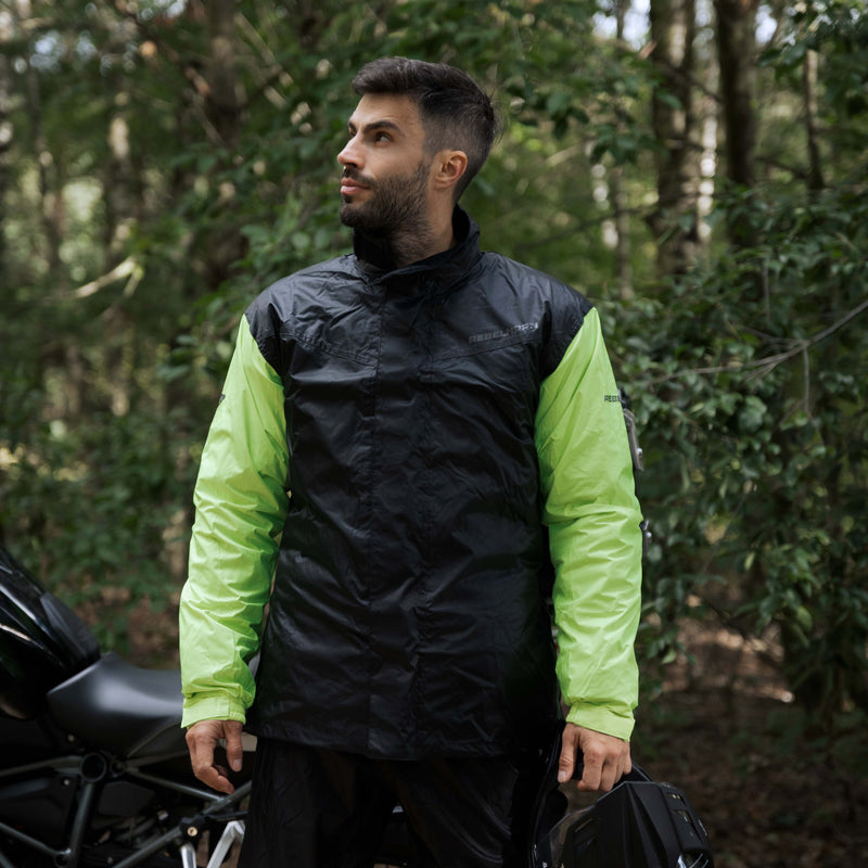 Man som står i skogen och bär Rebelhorn Regnställ 2-delat Rain Svart/Flu, idealiskt för skydd mot regn under motorcykelkörning.